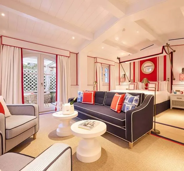 Quarto de hotel luminoso com paredes listradas brancas e vermelhas, uma cama grande com dossel, um sofá azul marinho com almofadas coloridas, duas mesas redondas brancas e portas de vidro que levam a um pátio.