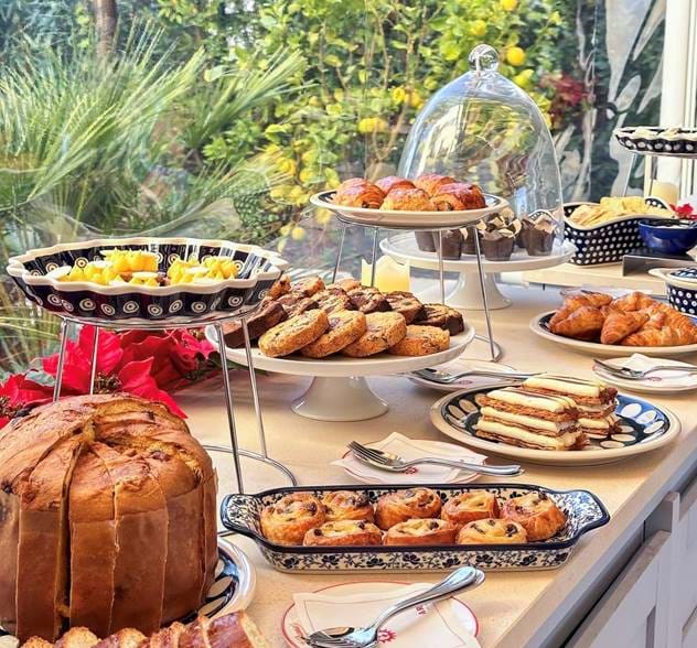 Set de table avec divers produits de boulangerie, notamment du panettone, des biscuits assortis, des pâtisseries, des croissants et des tranches de gâteau en couches, avec un jardin visible en arrière-plan.