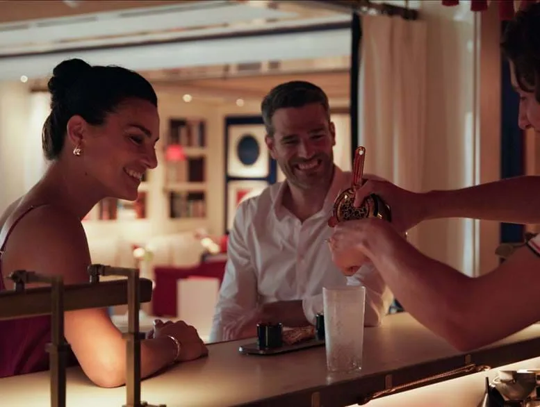 Casal sorridente sentado num balcão de bar enquanto o barman prepara um cocktail.