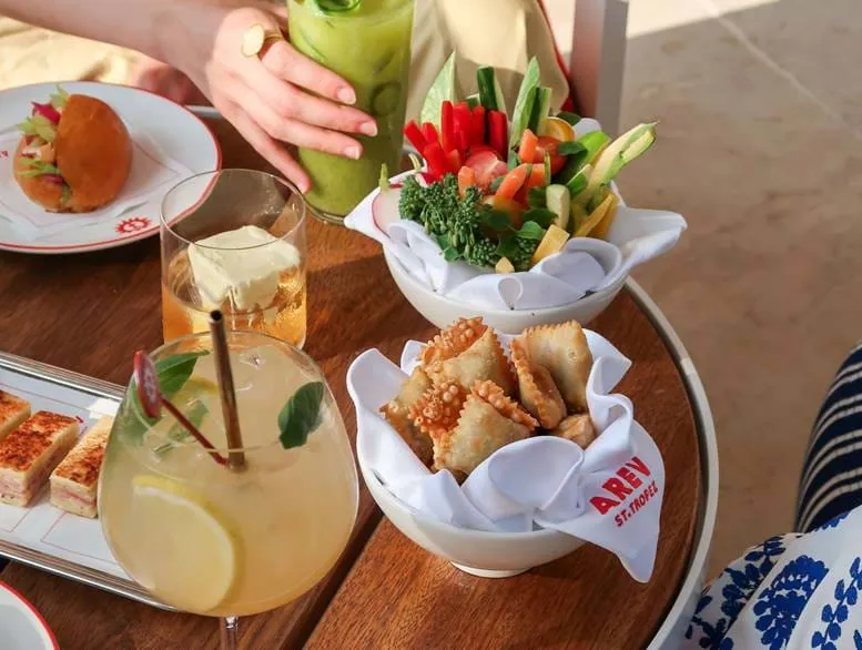 Tisch mit verschiedenen Vorspeisen, darunter gebratene Knödel in einer Schüssel, frische Gemüsesticks in einer Schüssel, ein Sandwich und drei verschiedene Getränke, von denen eines grün ist und von einer Person gehalten wird.