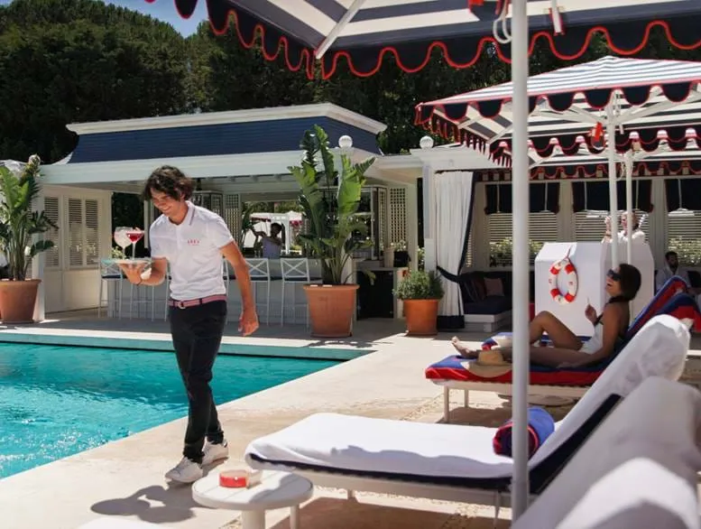 Ein Kellner mit Getränken geht neben einem Pool spazieren, an dem eine Frau auf einem Stuhl unter einem Sonnenschirm an einer Poolbar mit schattigen Sitzgelegenheiten liegt.