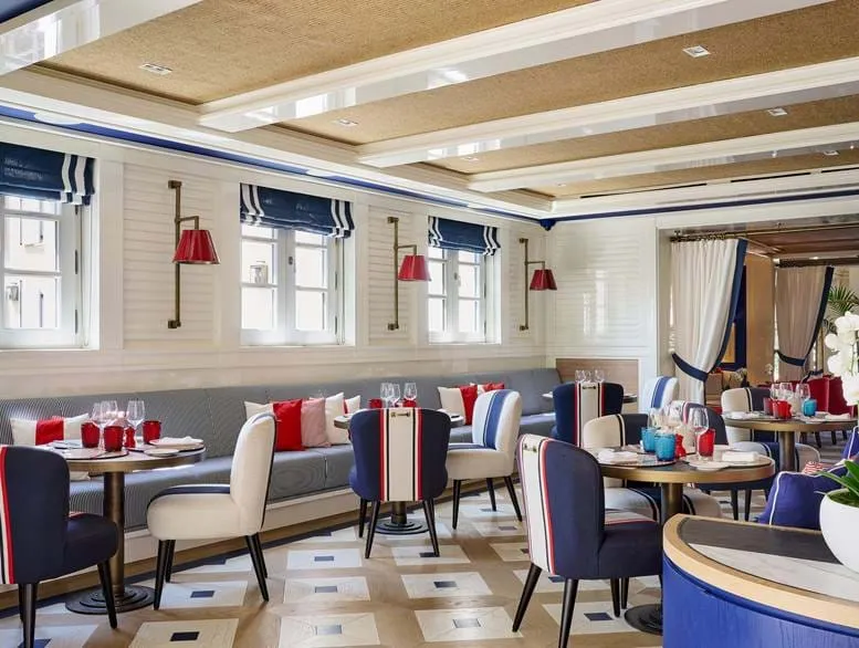 Elegante Lounge-Bar mit marineblauen, weißen und roten Stühlen, runden Tischen mit Gläsern und einer langen gepolsterten Bank unter Fenstern mit marineblau-weiß gestreiften Vorhängen.