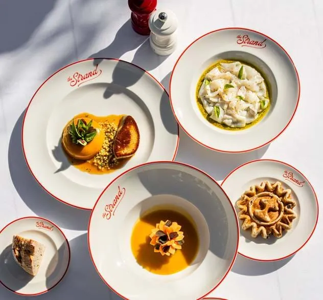 Fünf weiße Teller mit roten Rändern von The Strand, jeweils mit einem anderen Gourmetgericht, darunter ein Soufflé mit Beilage, Weißfisch mit grünen Kräutern, ein mehrschichtiges Dessert, eine kleine Torte und eine Scheibe Brot auf einer weißen Tischdecke mit Salz- und Pfefferstreuern.