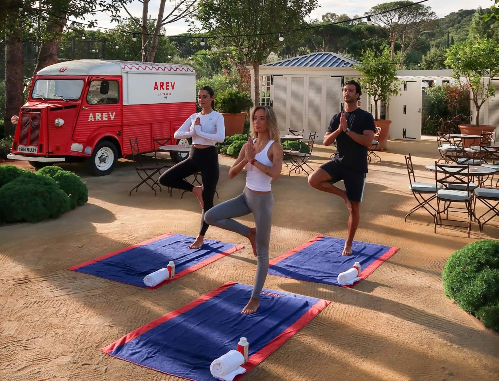 Aula de ioga ao ar livre nos jardins do hotel AREV em Saint-Tropez com uma van vintage ao fundo.