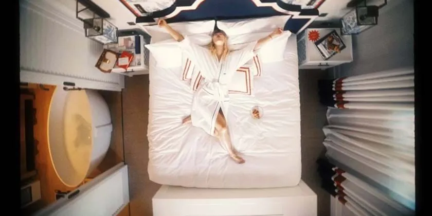 Vista de alto ângulo para um quarto de luxo no hotel AREV em Saint-Tropez, com uma pessoa relaxando numa grande cama branca com roupa de cama elegante, rodeada por um design interior elegante e luminoso.