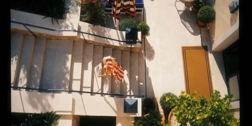 Vista ensolarada da fachada do hotel AREV em Saint-Tropez. Uma pessoa vestindo uma roupa laranja brilhante fica em uma varanda do primeiro andar, cercada por vegetação e elegante arquitetura mediterrânea.