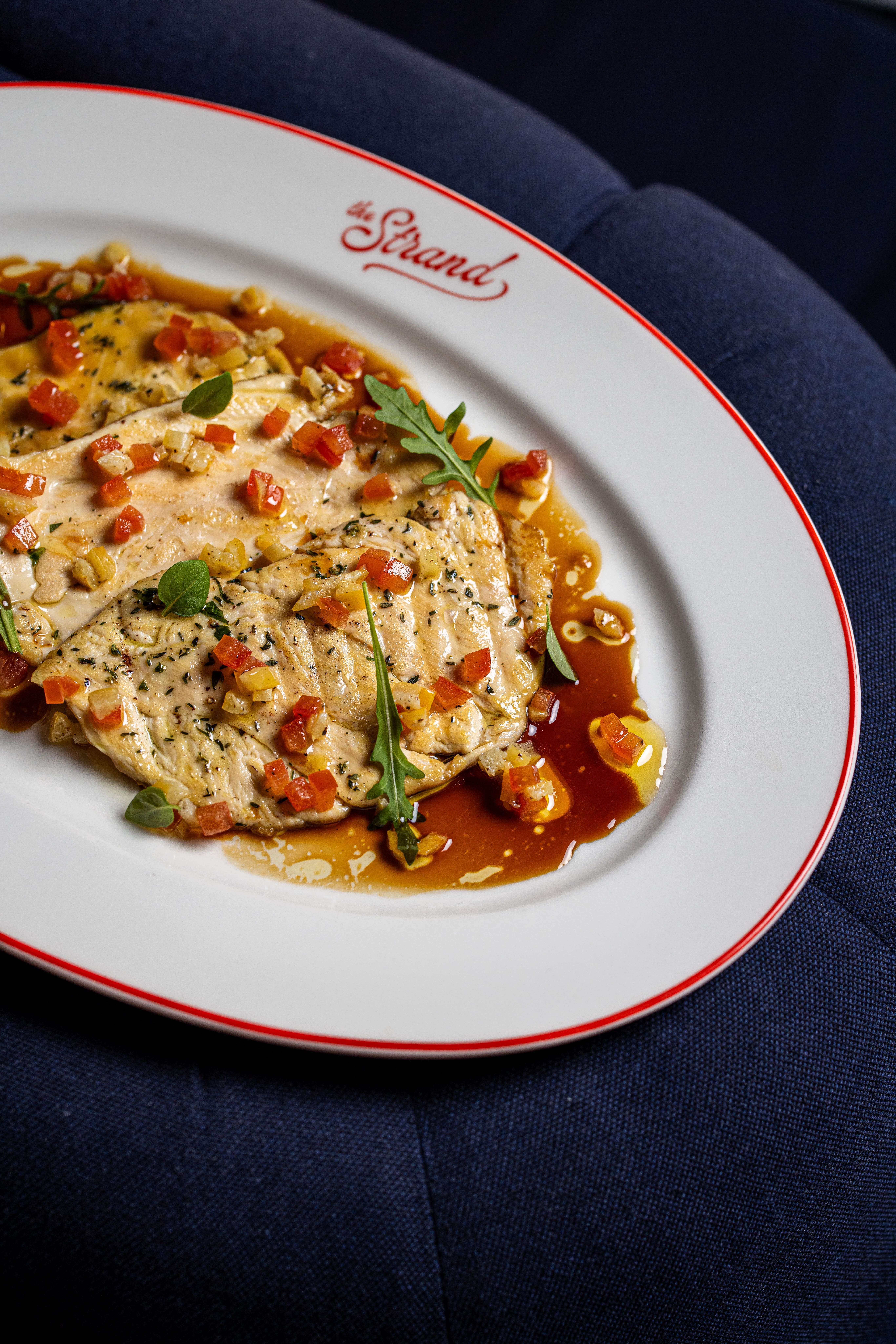 Filetes de peixe grelhados guarnecidos com legumes e ervas vermelhas e amarelos em cubos, servidos com molho castanho num prato branco com guarnição vermelha e logo 'The Strand'.
