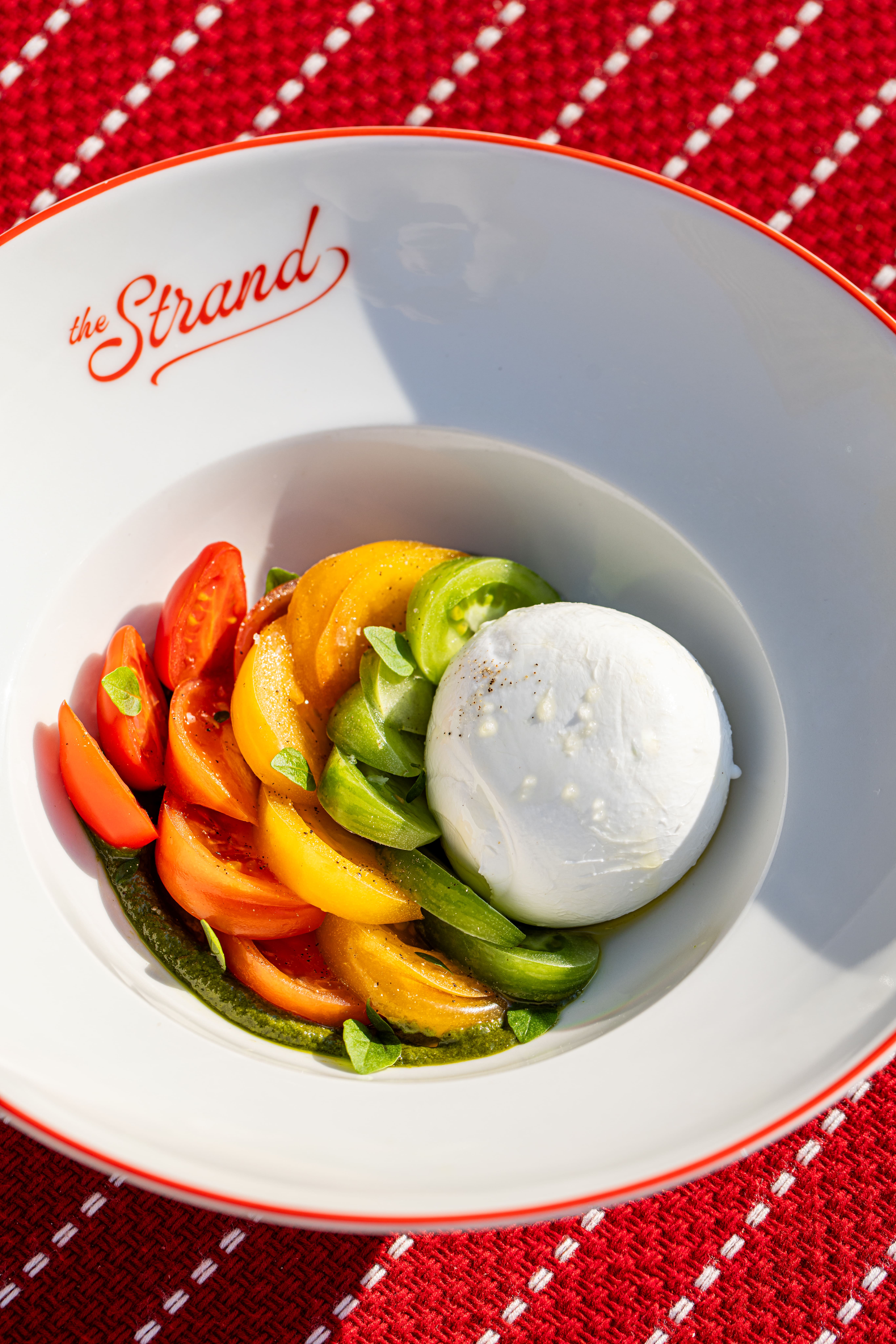 Um prato de tomate fatiado multicolorido e folhas de manjericão dispostos em torno de uma bola de queijo mussarela fresco com molho pesto num prato branco rotulado 'The Strand', sobre uma toalha de mesa estampada de vermelho.