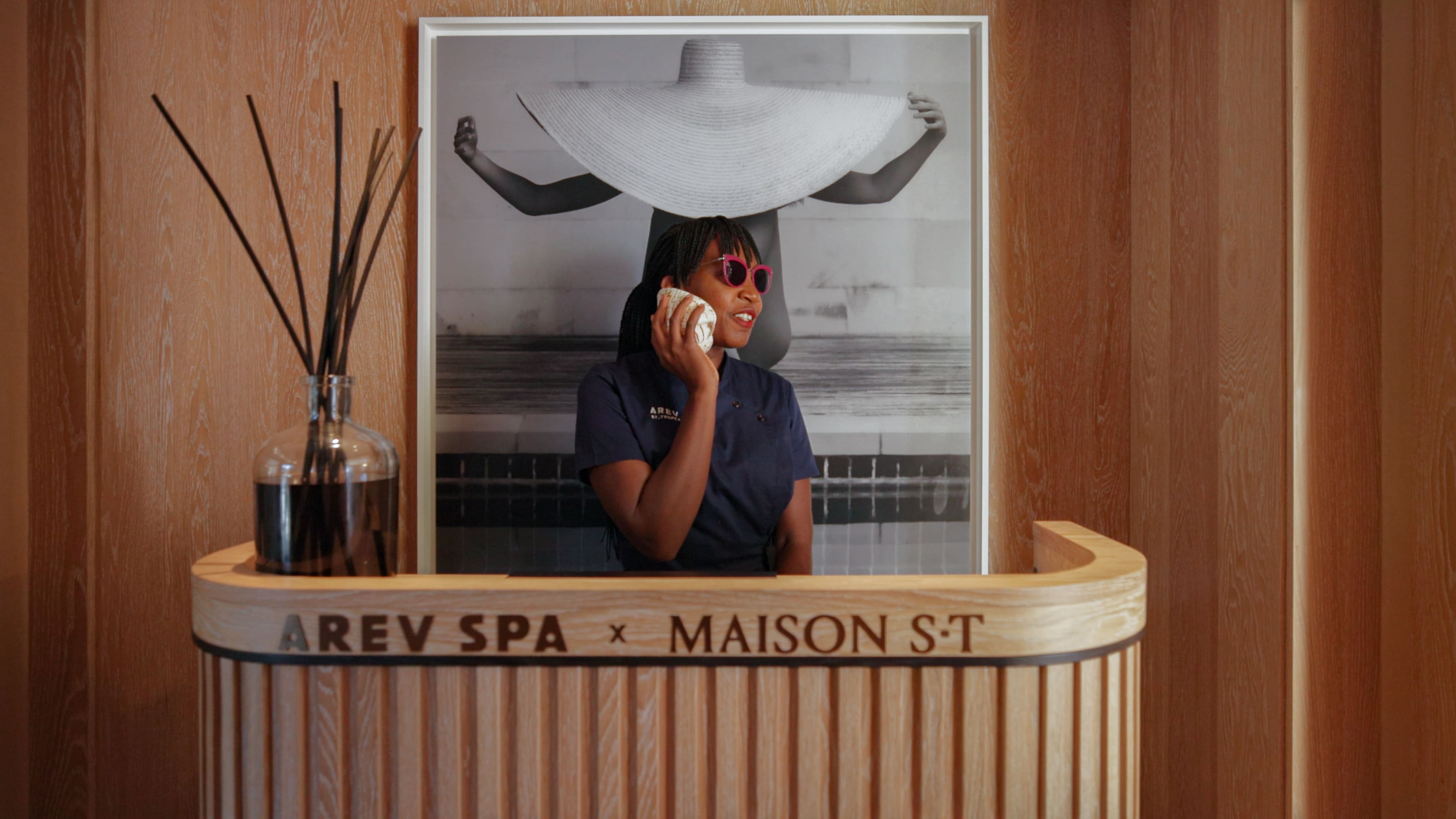 Mulher em uniforme de spa segurando uma concha na orelha, de pé atrás de uma recepção de madeira com texto 'AREV SPA x MAISON S.T', com uma grande foto em preto e branco de uma pessoa usando um chapéu largo ao fundo.