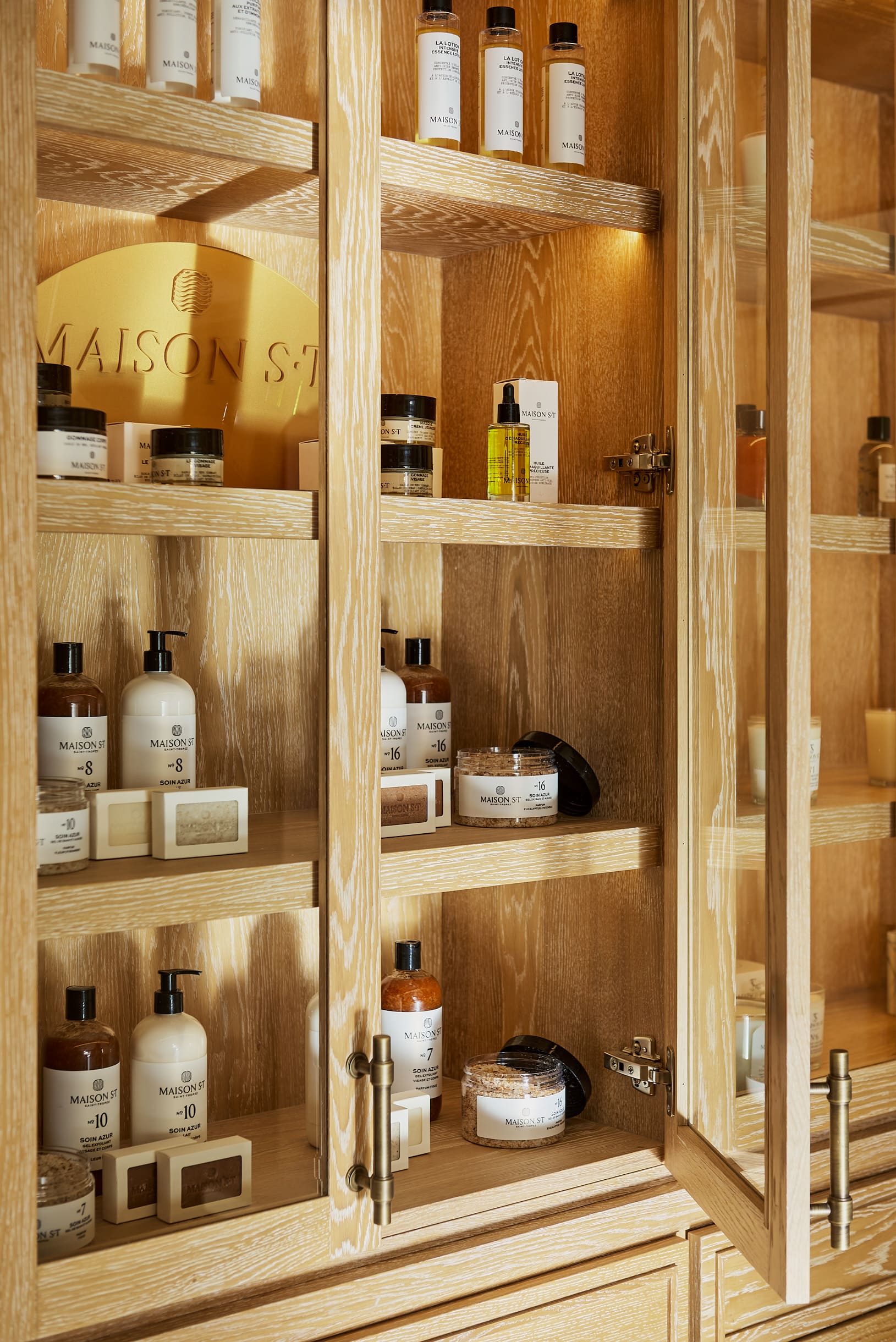Armário de madeira com portas de vidro exibindo produtos de beleza e produtos de beleza da Maison S.T. dispostos nas prateleiras.