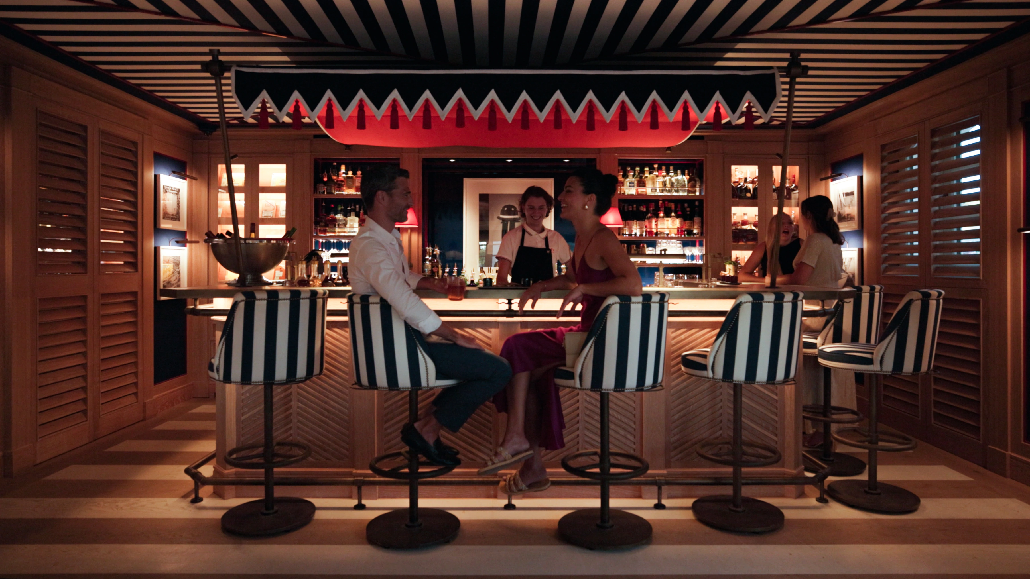 Pessoas desfrutando de bebidas e conversando em um bar elegante com cadeiras listradas e painéis de madeira.