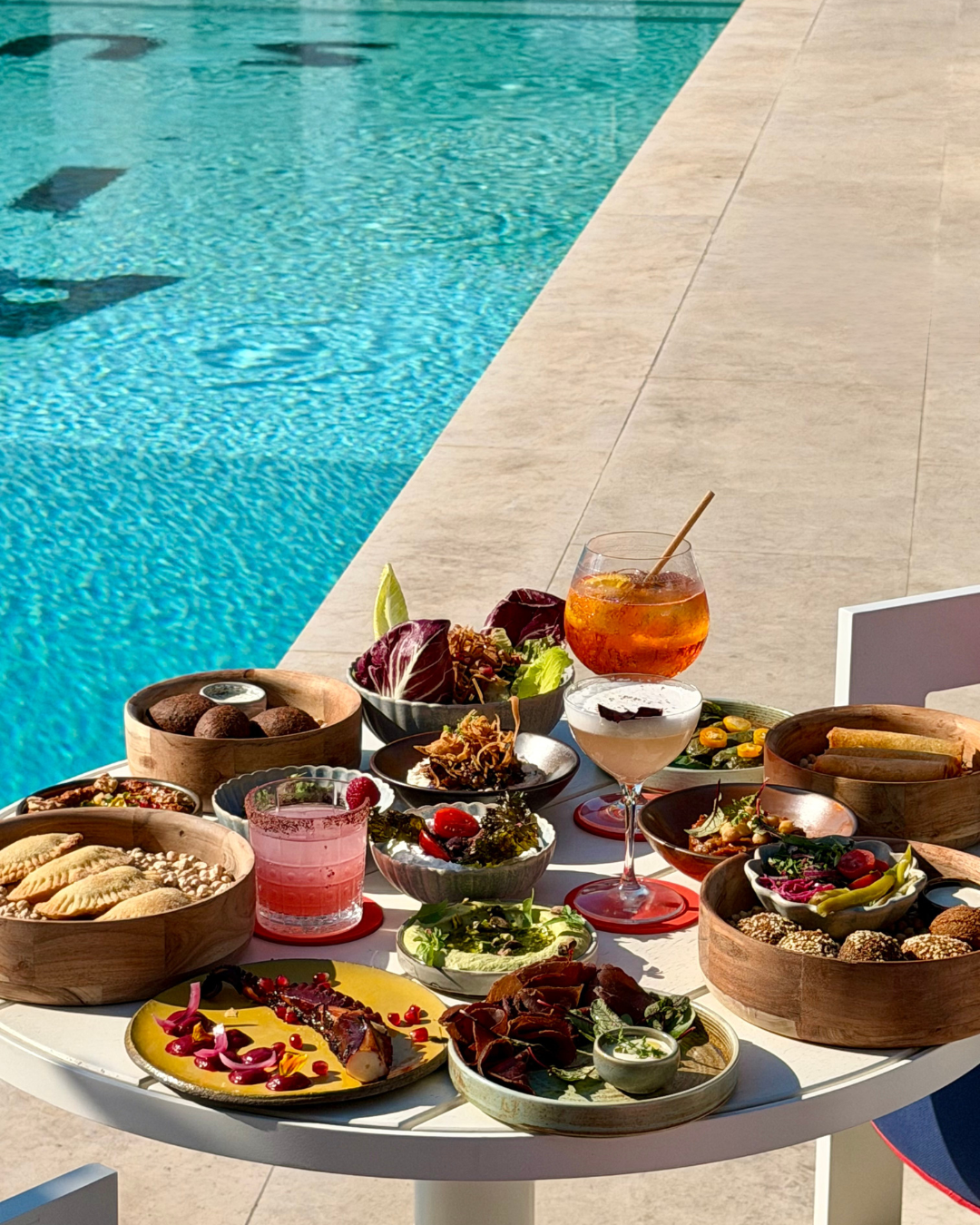 Outdoor table by pool with assorted appetizers, salads, and cocktails in glassware under sunlight.