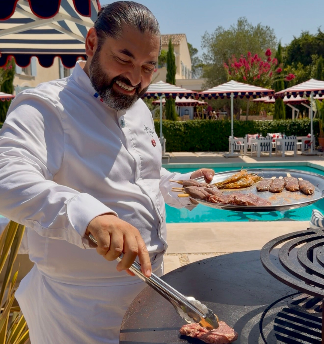 Chef grilling meat skewers outdoors by a pool with umbrellas and seating in the background.