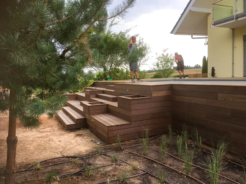 Zwei Männer arbeiten auf einer großen Holzterrasse mit Stufen vor einem gelben Haus.