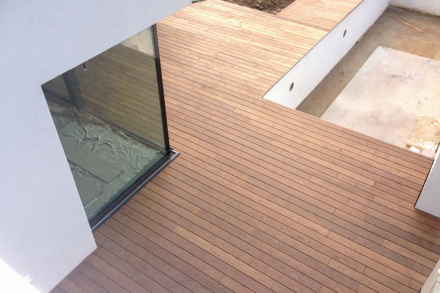 Moderner Balkon mit Holzterrasse und großem Glasfenster, angrenzend an einen ungefüllten Poolbereich.