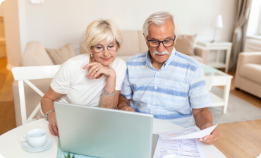 Senior couple sitting at a table, reviewing documents and using a laptop in a cozy living room.