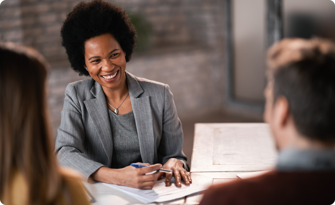 Smiling businesswoman in a gray suit holding a pen and talking to two people across a table in a professional setting.