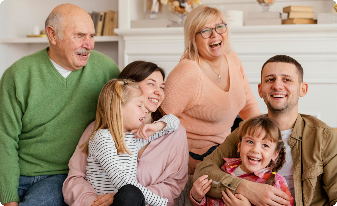 Three generations of a smiling family sitting together indoors, including a grandfather in a green sweater, a grandmother in glasses and peach sweater, and parents holding two young girls.