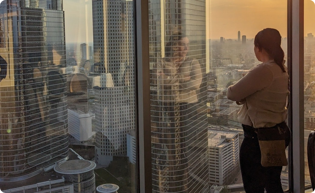 Person standing inside a high-rise building looking out over a city skyline at sunset.