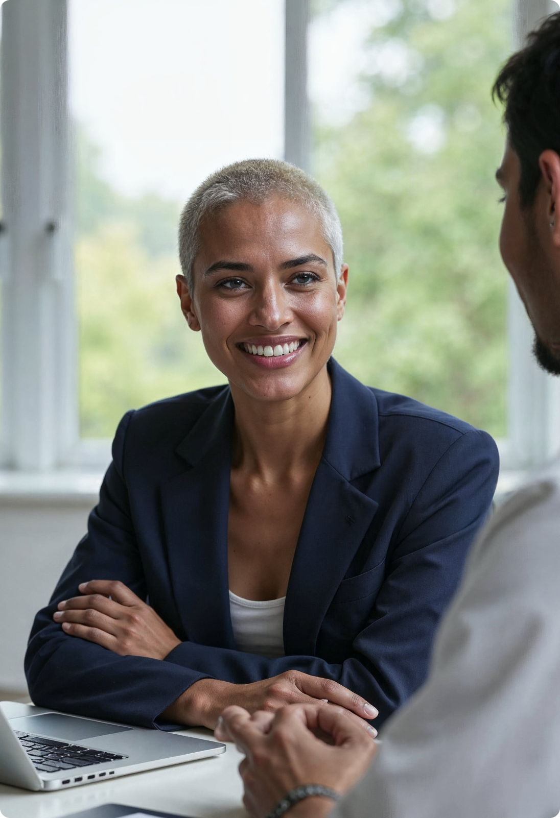 Smiling woman in a navy blazer engaged in conversation with a man across a table with a laptop.