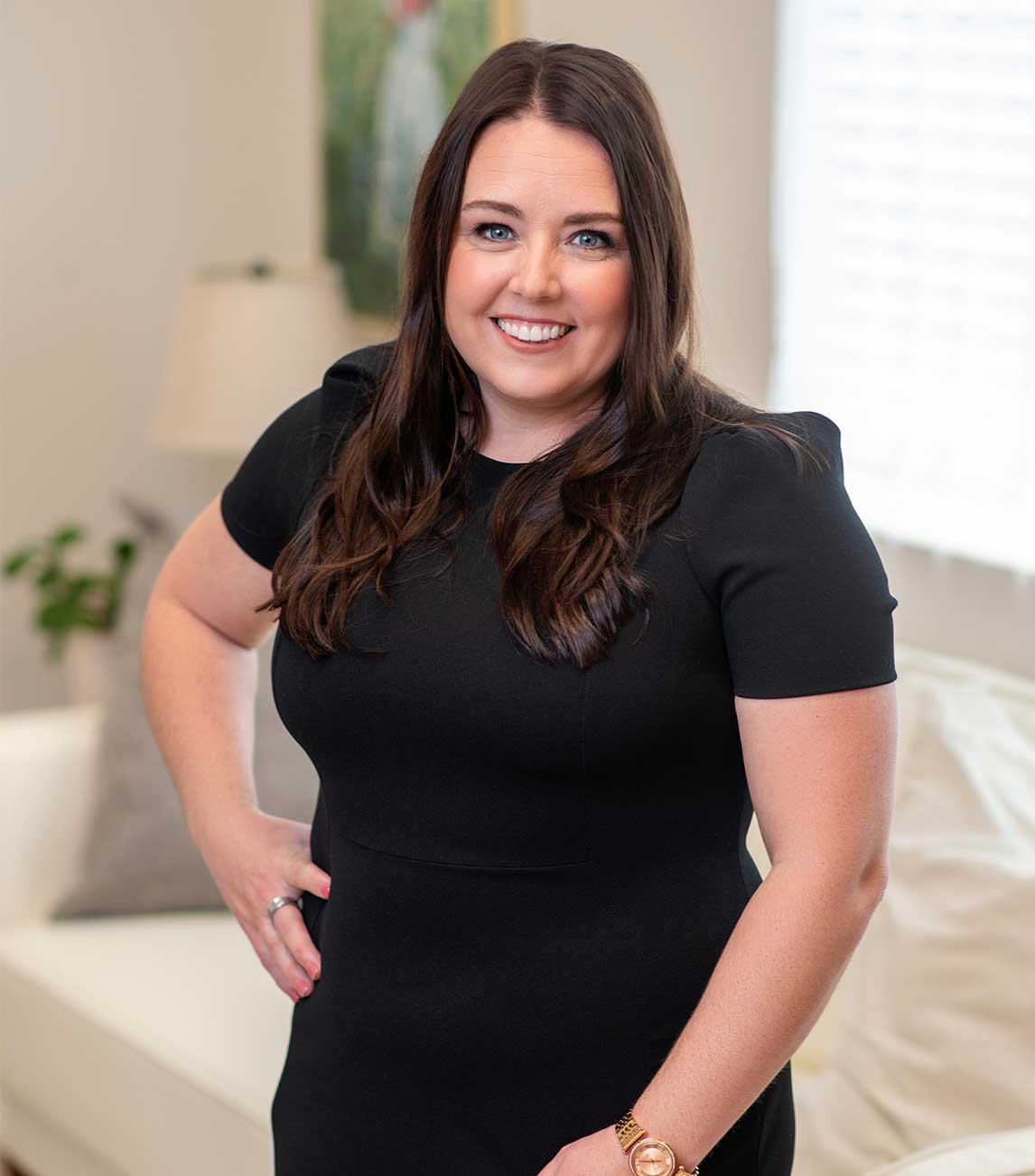 Smiling woman with long brown hair wearing a black dress standing indoors with a hand on her hip.