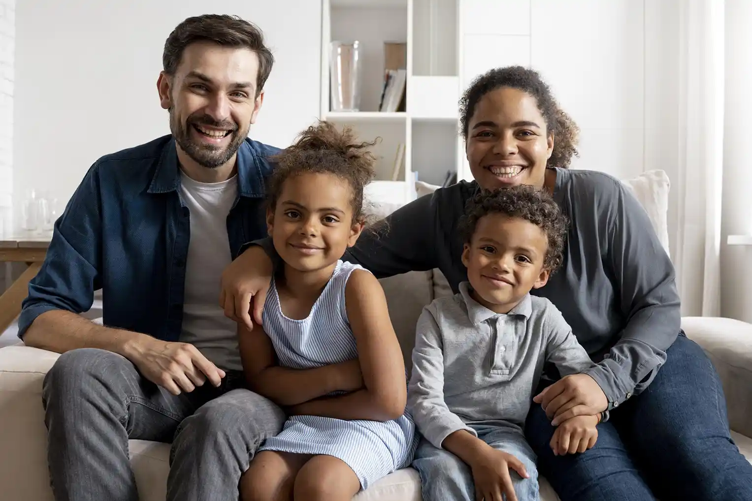 Smiling family of four sitting closely together on a couch in a cozy living room.