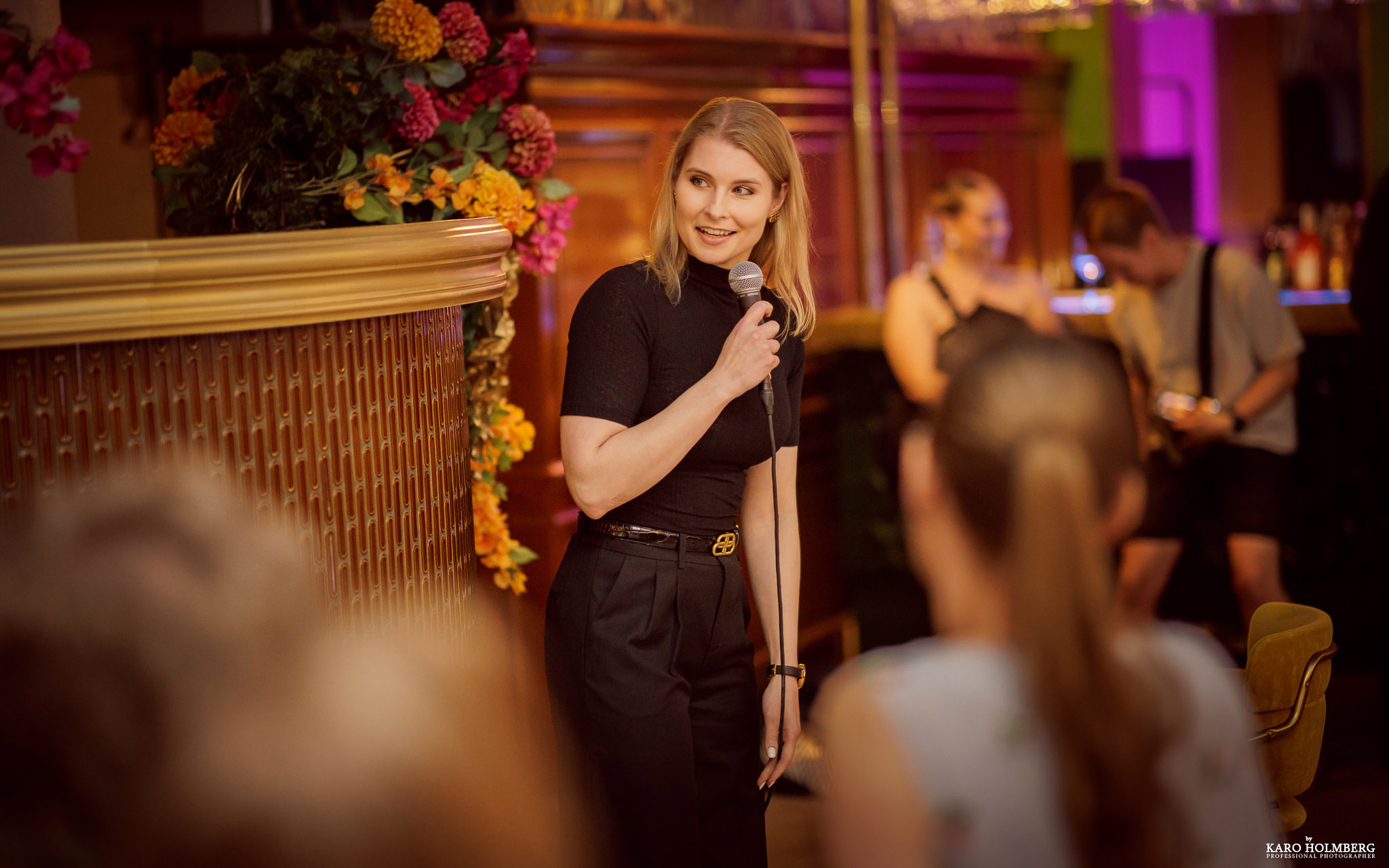 Stand Up -koomikko Taina Törmänen esiitymässä keikalla, taustalla ihmisiä ja kukka-asetelma.