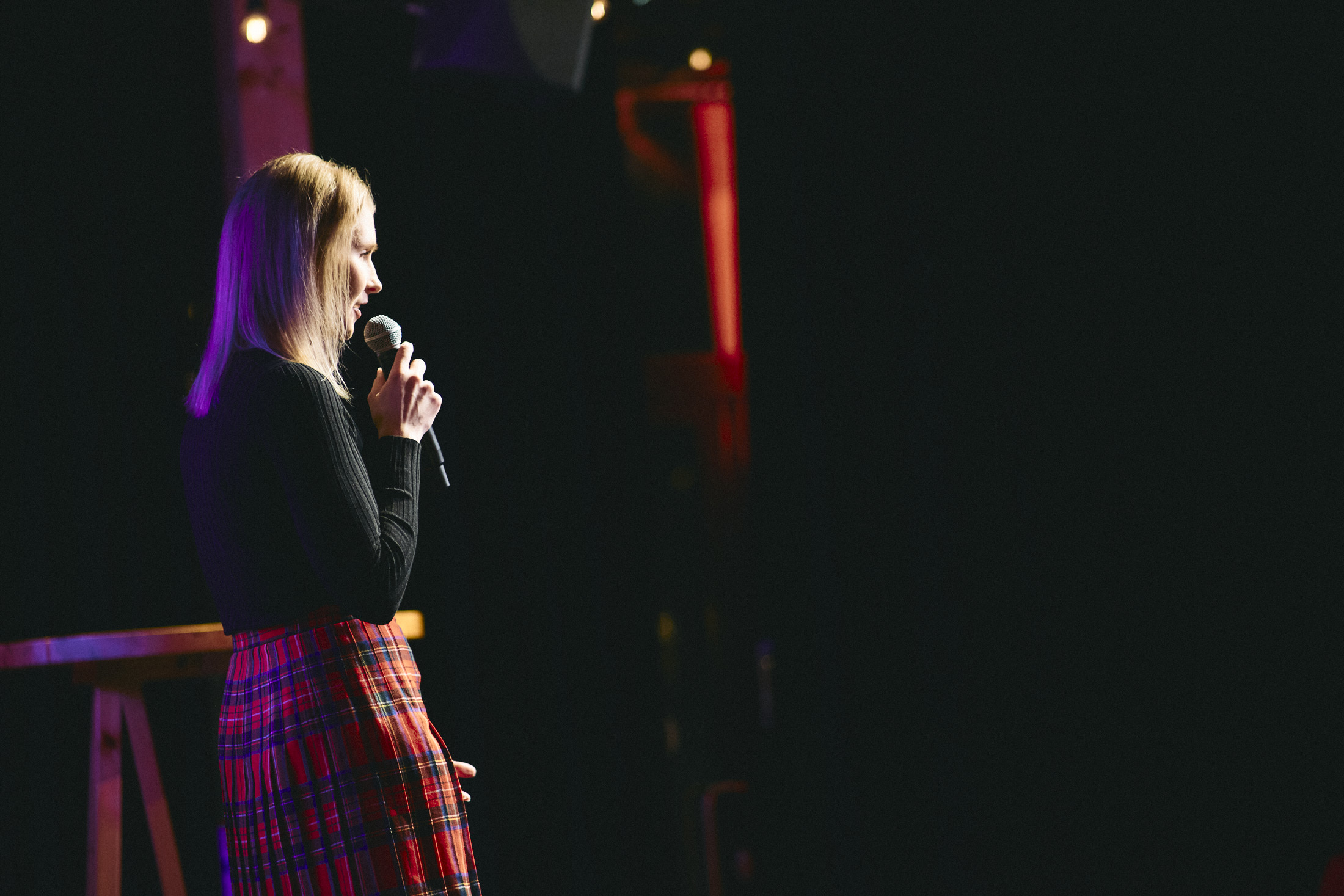 Stand Up -koomikko Taina Törmänen mikrofoni kädessä näyttämöllä tummaa taustaa vasten.