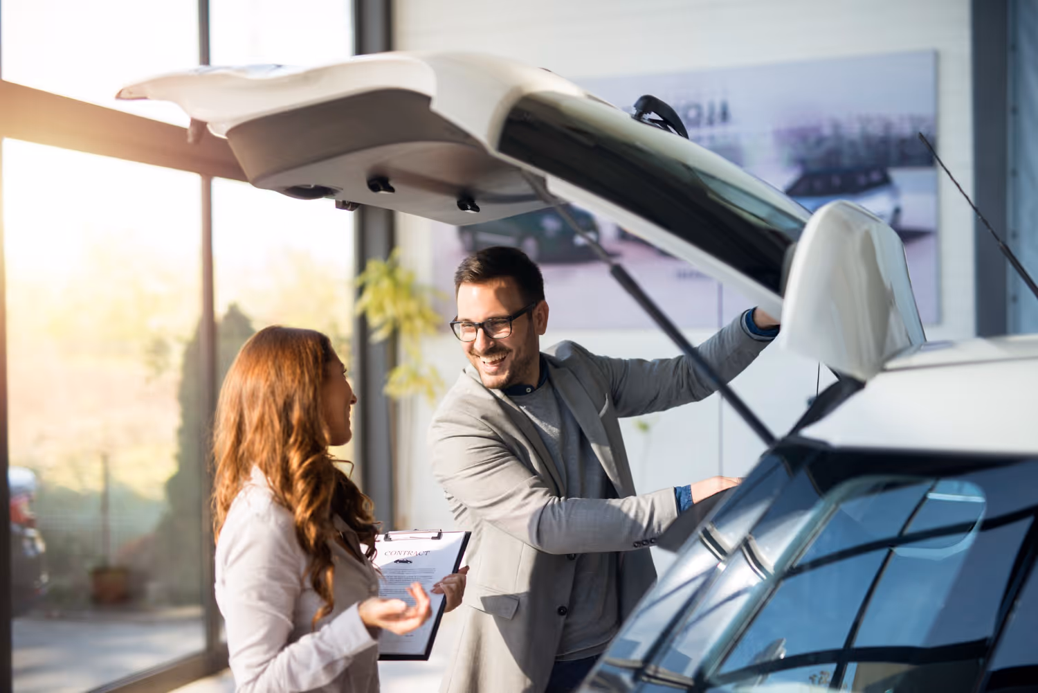 Man and woman smiling while examining the open trunk of a car in a bright showroom.