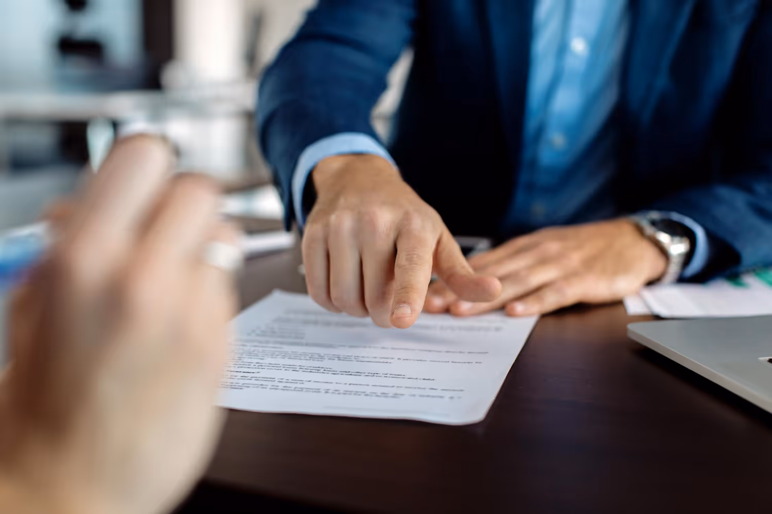 A person in a blue suit points at a document on a table during a discussion with another individual.