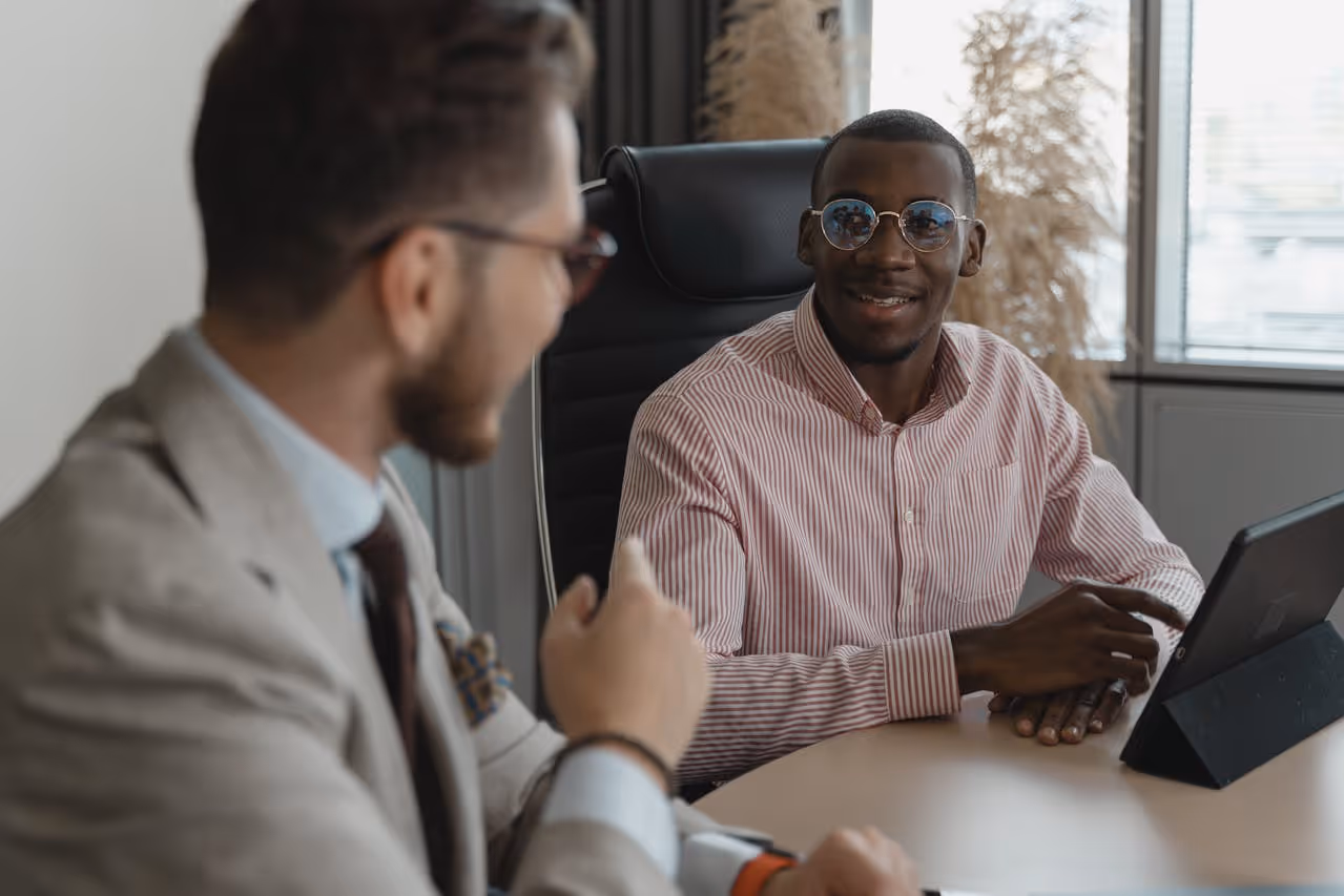 Two men having a discussion at a table, one wearing glasses and a striped shirt using a tablet.