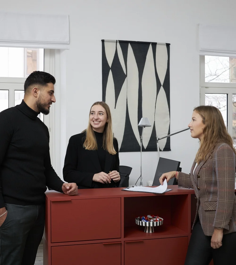 Drei junge Erwachsene sprechen an einem roten Empfangstresen in einem modernen Büro.