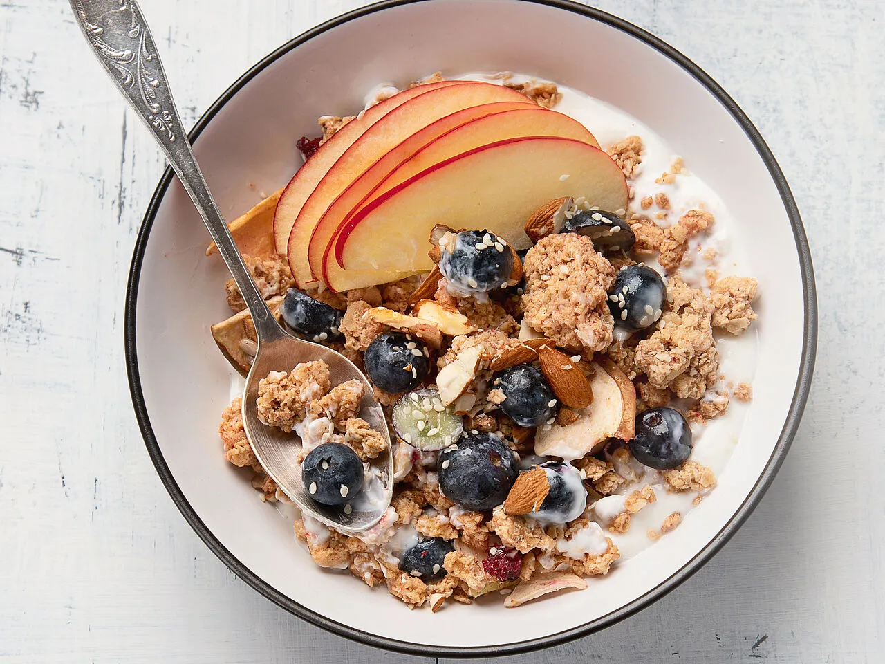 Schale mit Müsli, Apfelscheiben, Blaubeeren, Mandeln und Joghurt, Löffel darin.
