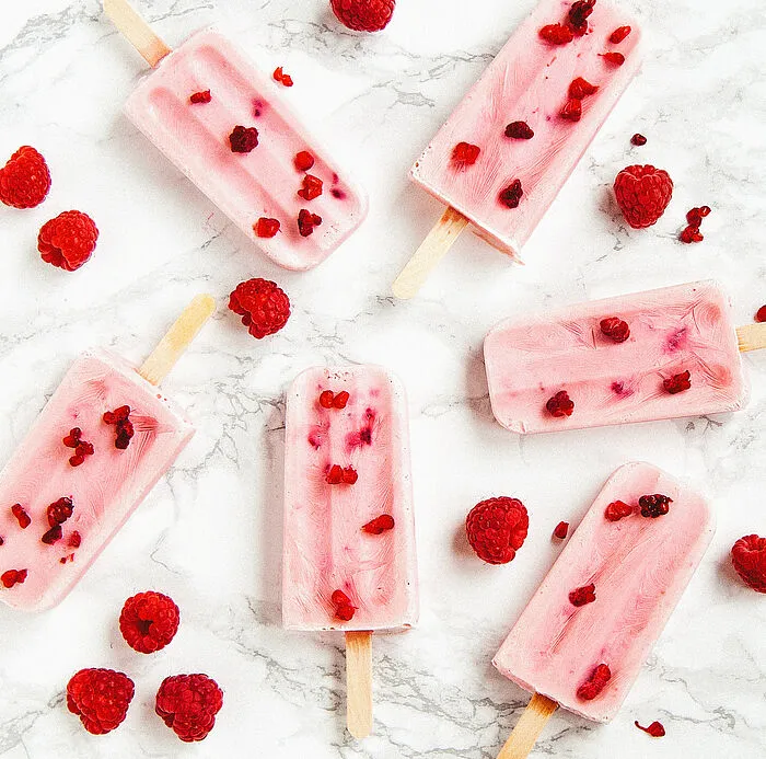 Sechs rosa Eis am Stiel mit Himbeerstücken und ganze Himbeeren auf weißem Marmor.
