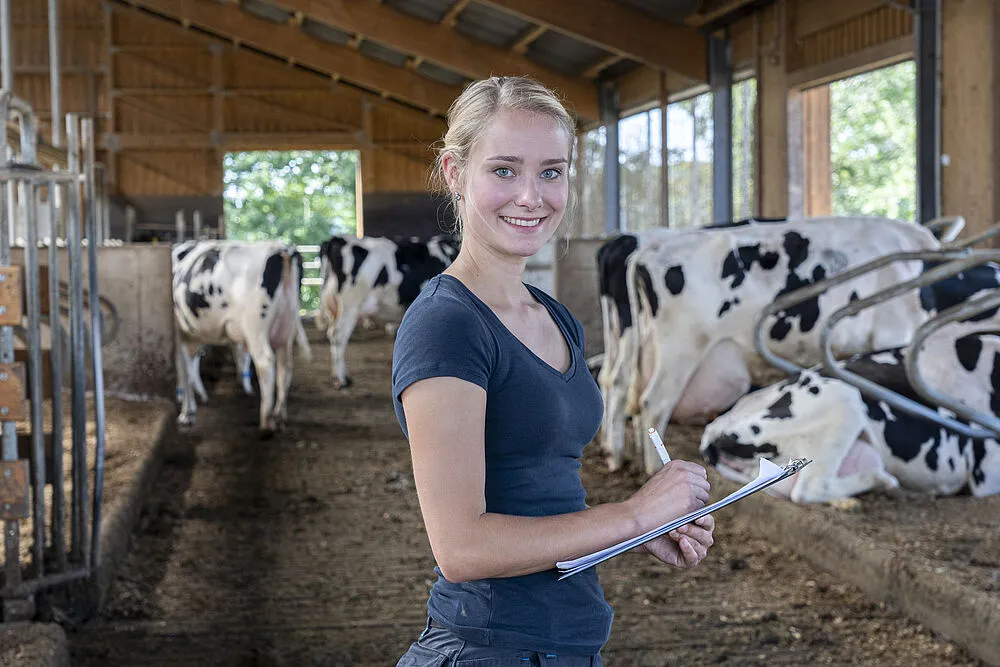Lächelnde Landwirtin mit Clipboard in einem Kuhstall mit schwarz-weißen Kühen.