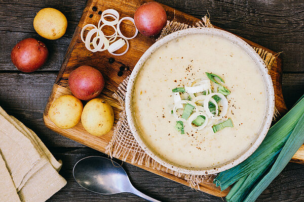 Schüssel mit cremiger Kartoffelsuppe, dekoriert mit Lauchringen auf Holzbrett mit Kartoffeln und Lauch.