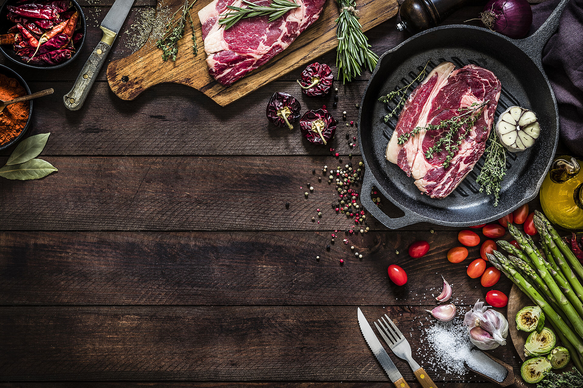 Rohes Rindersteak mit Kräutern in Pfanne und Zutaten wie Spargel, Tomaten, Knoblauch, Pfeffer auf Holz.