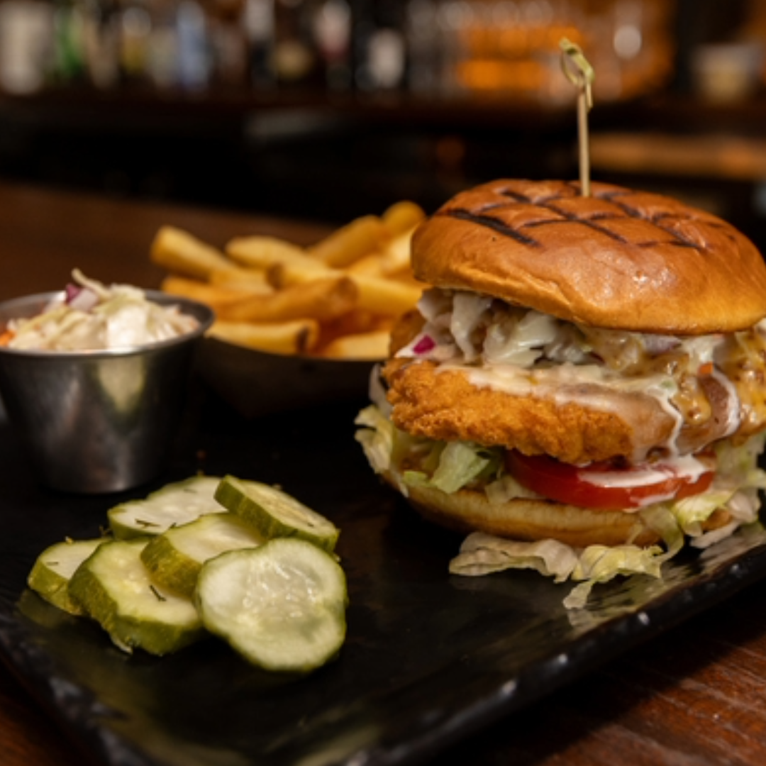 Fried chicken sandwich with lettuce, tomato, and sauce on a grilled bun served with pickles, coleslaw, and French fries on a black plate.