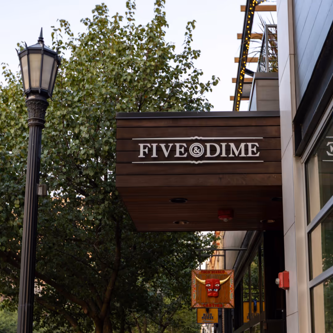 Street view of a brown wooden sign reading 'FIVE & DIME' mounted on a building with trees and a vintage street lamp nearby.
