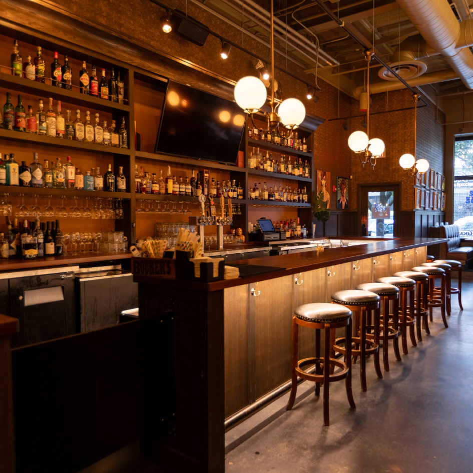 Interior of a warmly lit bar with a long wooden counter, cushioned bar stools, shelves with bottles and glasses, pendant lights, and a large TV mounted on the wall.