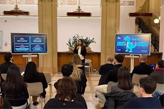 Evento digitale con relatore che parla davanti a un pubblico seduto, con due schermi che mostrano contenuti digitali in una sala storica.