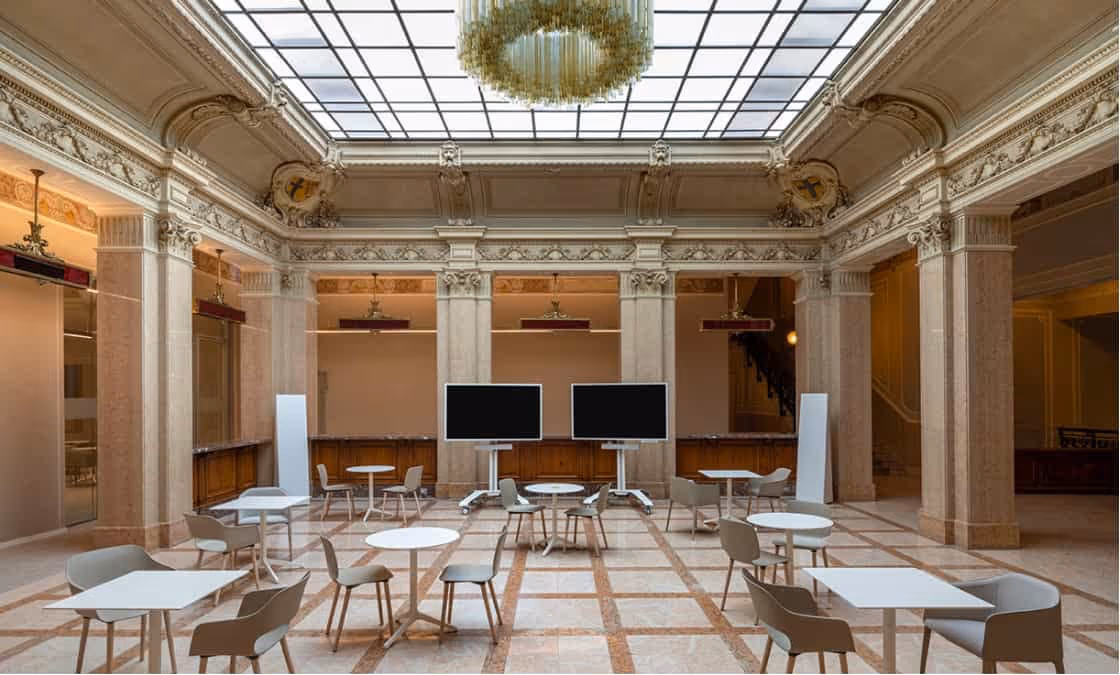 Spazio interno elegante con soffitto a vetri, grandi colonne decorati e tavoli bianchi con sedie moderne.