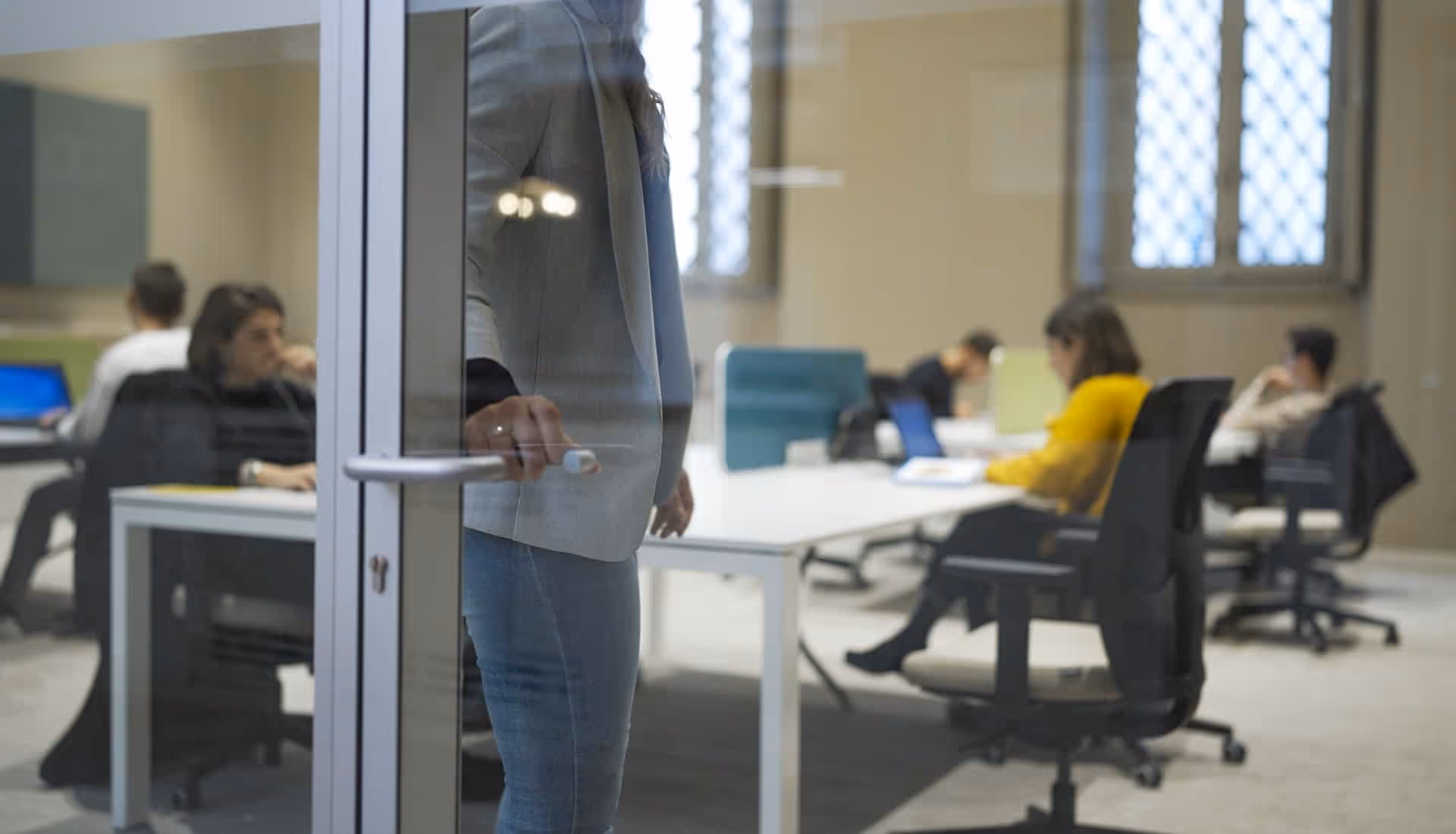Una persona apre la porta di vetro di un ufficio dove altre quattro persone lavorano ai loro laptop sedute a un tavolo bianco.
