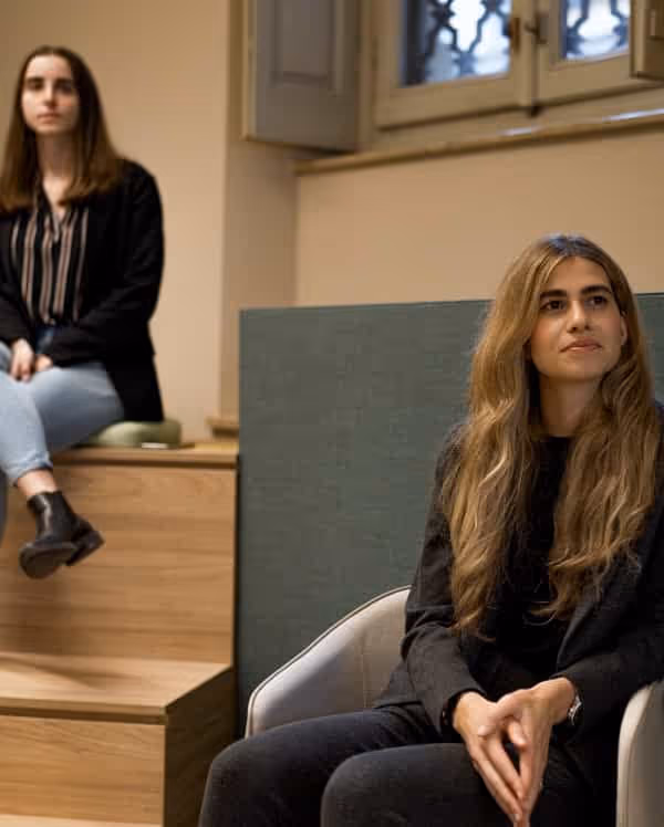 Due donne sedute in una stanza moderna, una su una poltrona chiara e l'altra su gradini in legno vicino a una finestra.