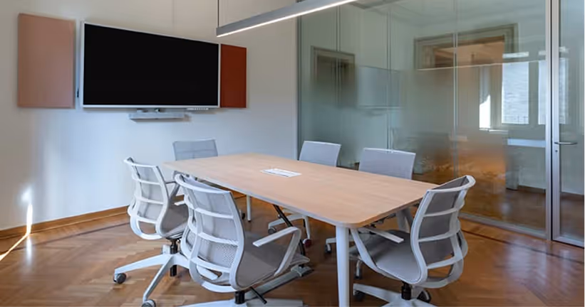 Sala riunioni moderna con tavolo in legno chiaro, sei sedie girevoli grigie e schermo TV a muro.