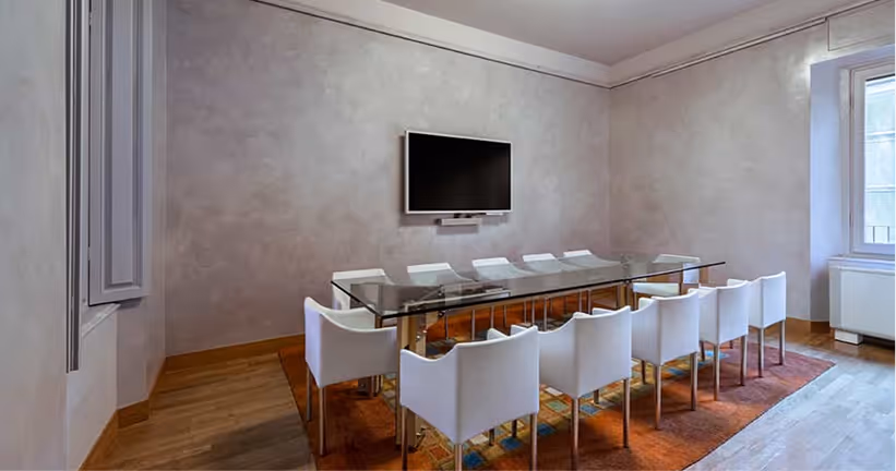Sala riunioni luminosa con tavolo in vetro, dieci sedie bianche e televisore a schermo piatto montato sulla parete.