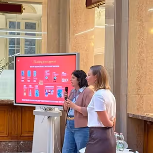 Due donne in piedi che tengono un microfono mentre presentano davanti a uno schermo con grafici rossi in una stanza con pareti in marmo.