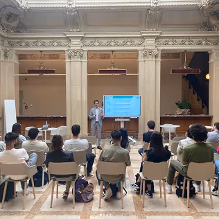 Relatore accanto a uno schermo con presentazione in sala conferenze con architettura classica, pubblico seduto volto verso il relatore.