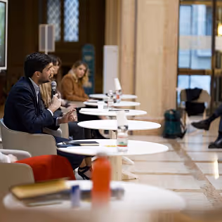 Persone sedute in una conferenza mentre un uomo parla al microfono in un ambiente con tavoli bianchi tondi.