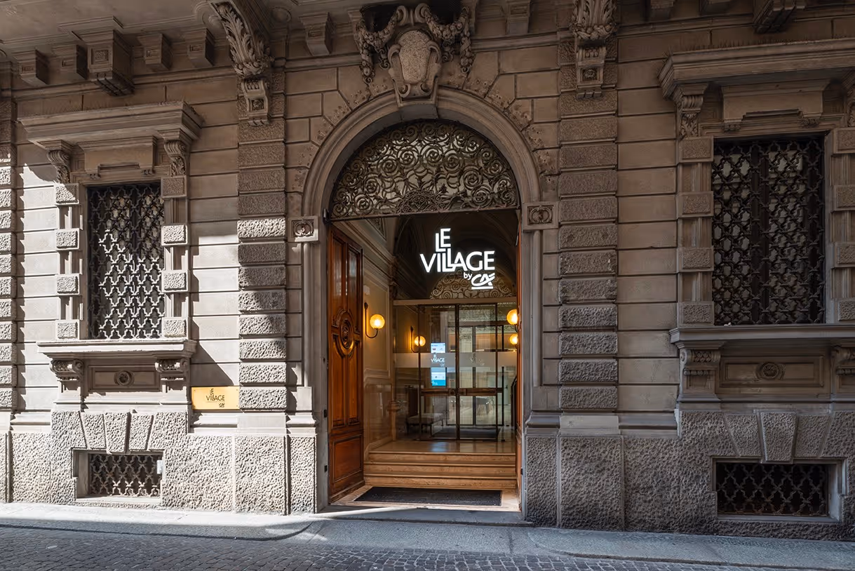 Ingresso storico con porta in legno aperta e insegna luminosa Le Village by CA sopra l'arco di pietra.